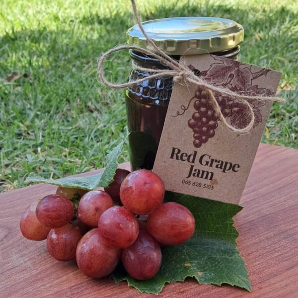 Homemade red grape jam in a glass jar with a golden lid, featuring deep red color and a rustic Anita’s Kitchen label, perfect for baked goods and pancakes.